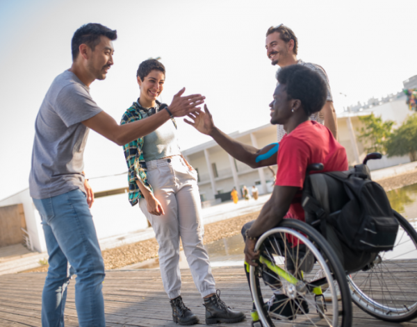 4 people are smiling and greeting each other. 3 stand, 1 is in a wheelchair
